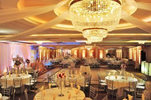 Luxurious quinceañera celebration at Astoria Banquets with a beautifully decorated gold and white ballroom and grand chandelier. Celebración de quinceañera de lujo en Astoria Banquets con un salón de baile hermosamente decorado en oro y blanco y un gran candelabro